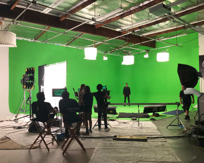 A film crew works on a green screen set with a lighting setup and several cameras aimed at a performer in the center of the studio.