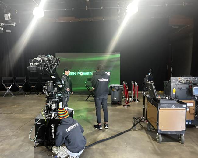 A television production studio is set up with a camera, crew members, and a backdrop displaying "GREEN POWERED."