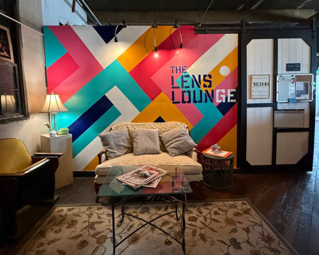 The image shows a cozy seating area in a space named "The Lens Lounge," featuring a vibrant, geometric mural and a glass coffee table.