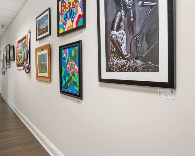 The image depicts a hallway lined with various framed artworks on the walls, showcasing colorful paintings and photographs.