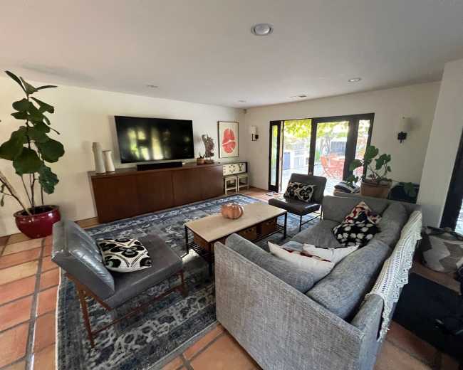 The image shows a modern living room with a television, a coffee table, and seating arranged around a patterned rug, featuring large windows that let in natural light.