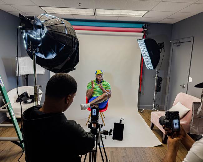 A man sits on a stool in front of a backdrop while being photographed by two individuals holding cameras in a well-lit studio.