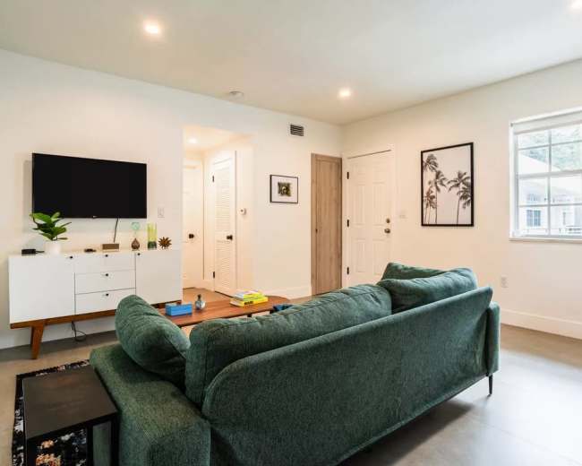 The scene features a modern living room with a green sofa, a television on a white media console, and a doorway leading to a hallway with light-colored walls.