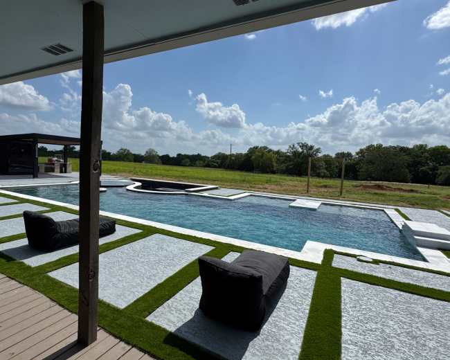 The image shows a modern swimming pool surrounded by landscaped grass and black bean bag chairs, with a lush green field and blue sky in the background.