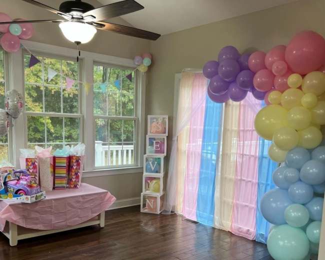 The room features a decorated corner with a table covered in a pink cloth, gifts stacked nearby, and a colorful balloon arch against the wall.