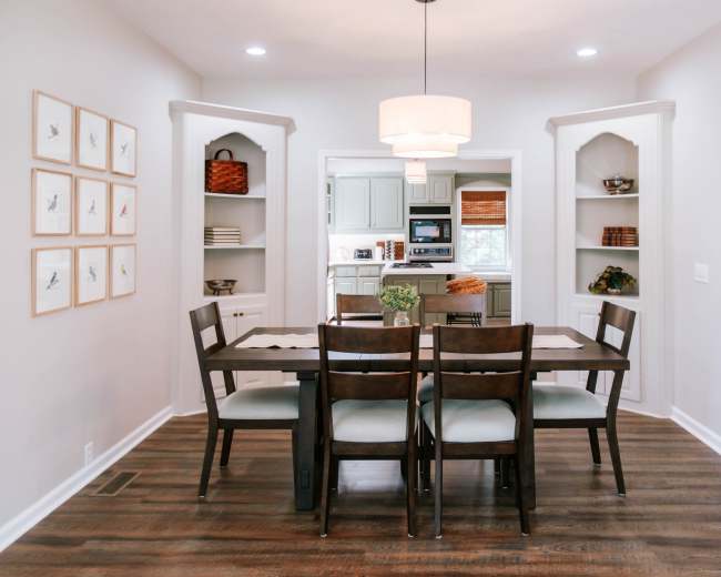 The image shows a well-lit dining area with a wooden table and chairs, framed bird prints on the wall, and open shelves displaying decorative items.