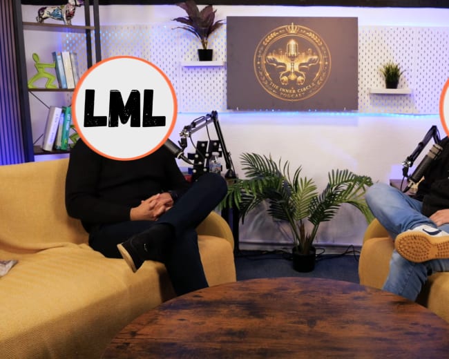Two individuals sit in yellow chairs across from each other at a round wooden table in a studio setting, with microphones positioned nearby and a plant in the background.