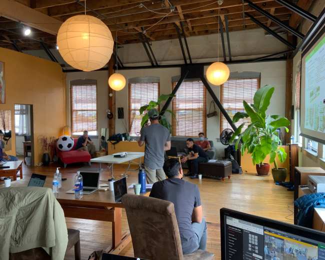 The scene shows a spacious room with large windows, various seating areas, and people engaging in discussions around a table, while plants and modern decor enhance the environment.