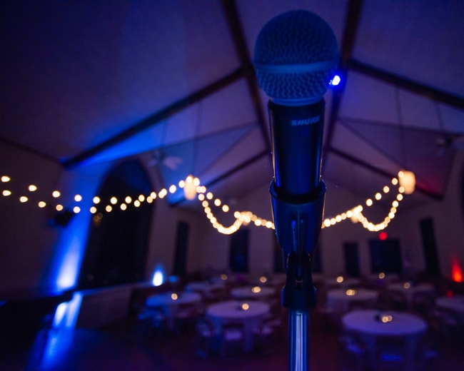A microphone stands in the foreground with a decorated venue featuring tables and string lights in the background.