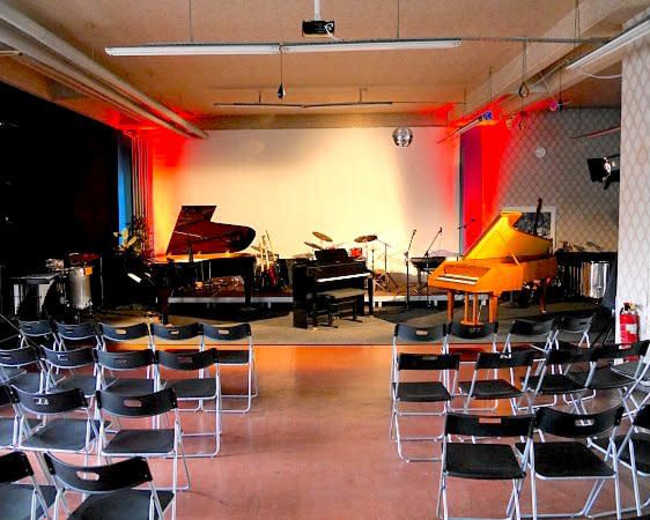 The image shows a music performance space with several pianos and a set of empty black chairs arranged in rows facing the stage.
