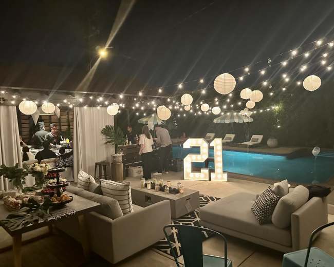 The scene depicts an outdoor party area at night, featuring a pool, string lights, and a large illuminated "21," with guests mingling and enjoying food and drinks.