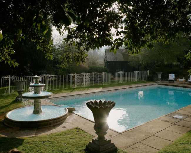 A clear swimming pool is enclosed by a white fence, featuring a fountain and decorative planters on the surrounding lawn.