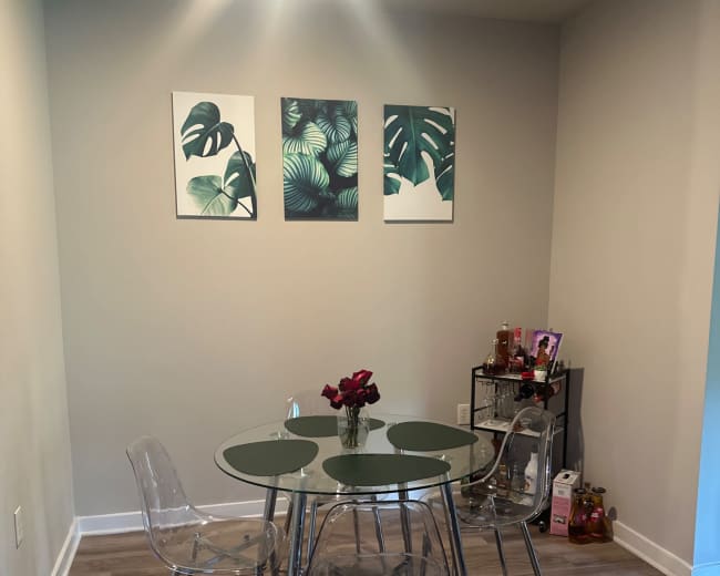 A glass-top dining table surrounded by four clear chairs is set against a wall decorated with three tropical-themed art pieces.