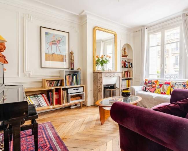 The image shows a stylish living room featuring a white sofa with colorful cushions, a purple armchair, bookshelves filled with books, and a large mirror above a decorative fireplace.