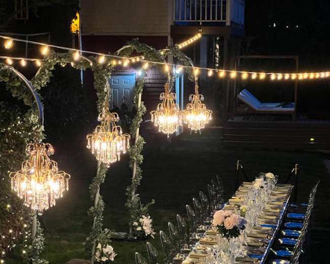 A long, elegantly set dinner table with crystal chandeliers and string lights is arranged outdoors, surrounded by greenery.