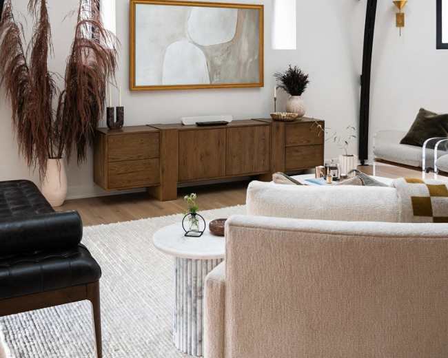 A modern living room features a light color palette, a large abstract painting on the wall, a dark leather sofa, and a mixture of decorative plants and furniture.