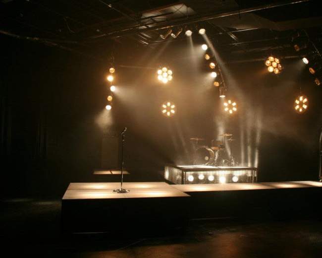 A dimly lit stage is set with spotlights, a microphone stand, and a drum kit surrounded by large amplifiers.