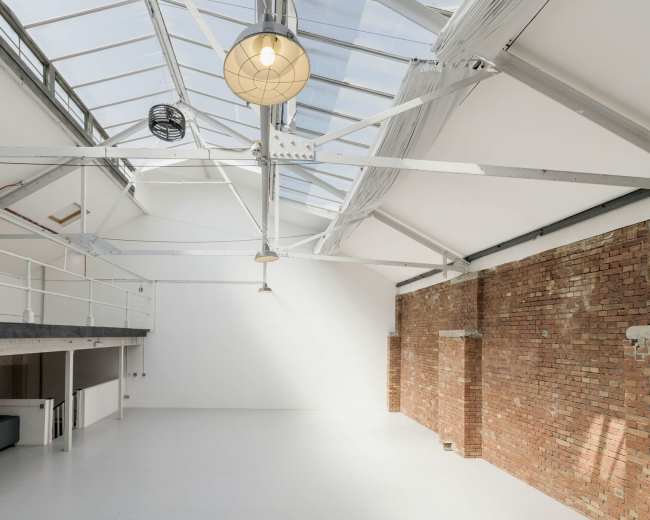 The image shows a bright, spacious industrial loft with a skylight, exposed brick wall, and metal beams.