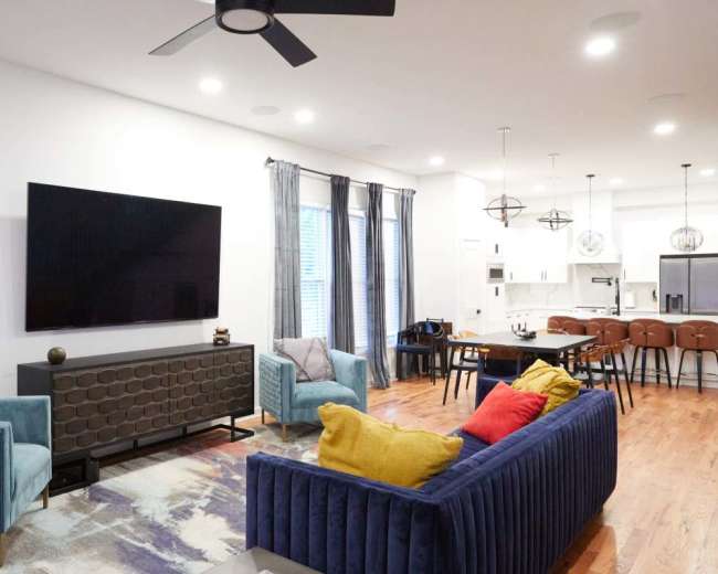 The image shows a modern living room with two upholstered sofas, a large wall-mounted television, a dining area with a table and chairs, and wood flooring.