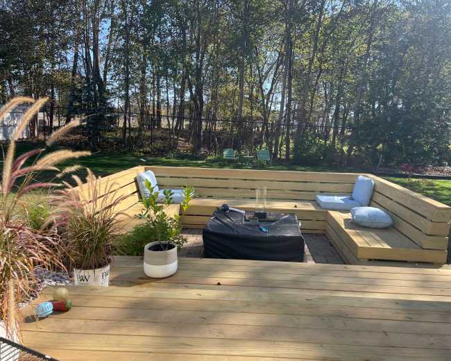 The image shows a wooden patio with a sectional seating area, a coffee table, and potted plants, all surrounded by trees in a sunlit outdoor environment.