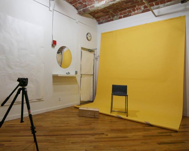 The image shows a photography studio with a yellow backdrop, a tripod with a camera, a chair, and various blank surfaces on the walls.