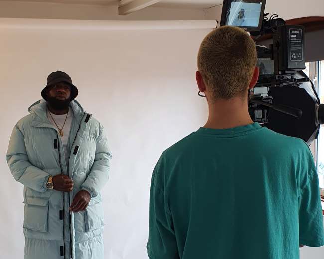 A man in a light blue puffer coat stands in front of a camera, while a crew member operates the equipment from behind.