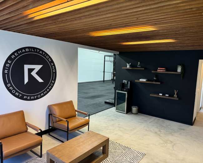 The image shows a modern interior of a rehabilitation and sports performance facility featuring brown leather chairs, a wooden coffee table, and black and white walls with a logo.