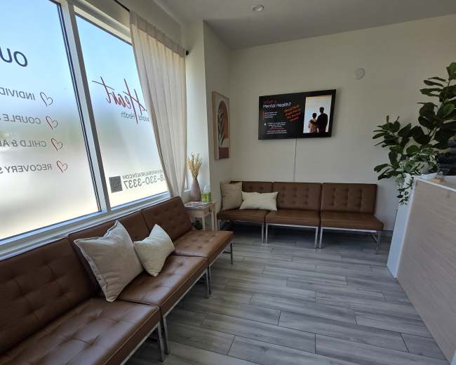 The image shows a modern waiting area with brown tufted couches, a light wooden reception desk, and large windows allowing natural light to enter.