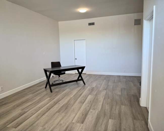 The image shows a sparsely furnished room with light-colored walls, a desk and chair, and laminate flooring.