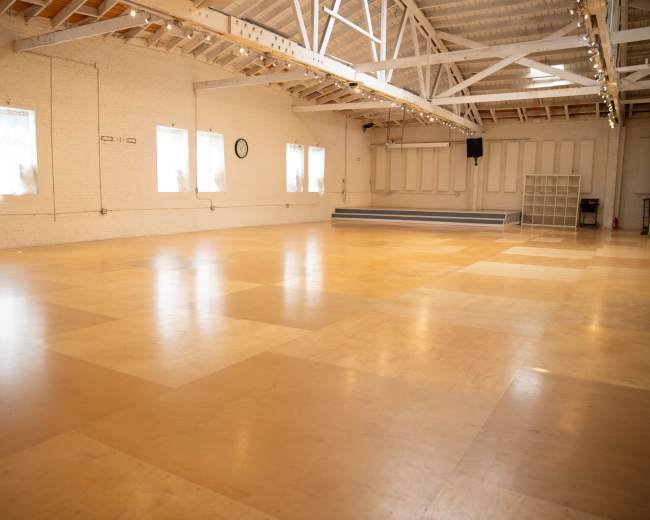 The image shows a spacious, empty studio with wooden flooring and large windows letting in natural light.