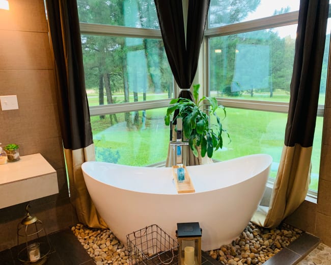 A freestanding bathtub is positioned in front of large windows that offer a view of a grassy area and trees outside.