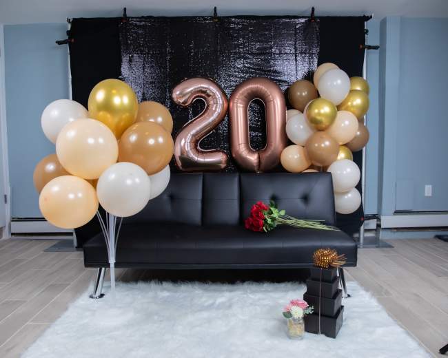 A black sofa is decorated with large metallic "20" balloons and an assortment of pastel balloons, alongside a bouquet of red roses and gift boxes.