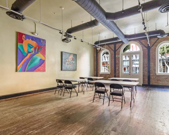 The image shows a spacious, vacant room with wooden floors, large arched windows, and several folding tables with chairs arranged in a row, decorated with colorful artworks on the walls.