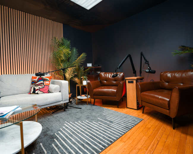 A cozy room features two brown leather armchairs, a gray sofa, a glass coffee table, and decorative plants against a dark wall.