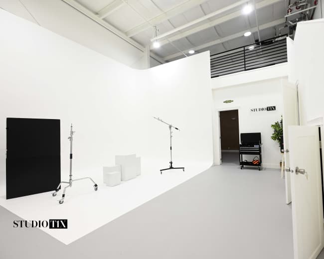 The image shows a spacious, well-lit studio with white walls, various equipment stands, and a door leading to another room.