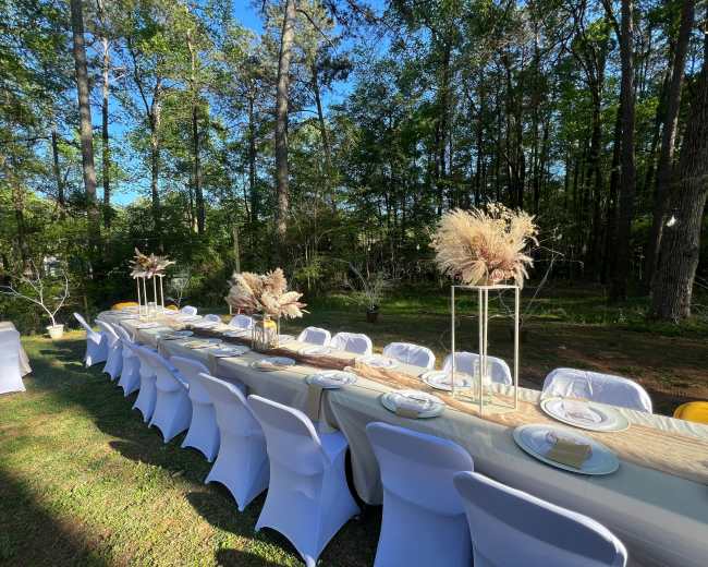 A long outdoor dining table is set with decor and chairs amidst a wooded area.