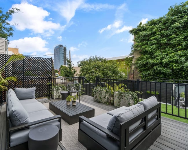 The image shows a modern outdoor terrace featuring gray seating, a coffee table, and various potted plants against a backdrop of a city skyline.