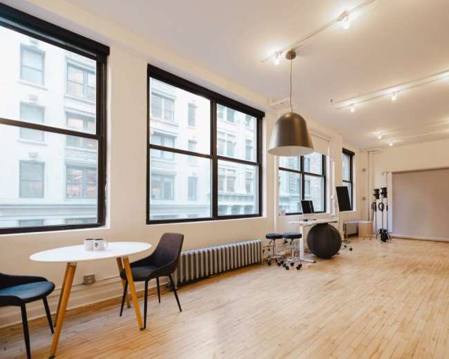 The image shows a well-lit, minimalistic studio space with large windows, a round table, two chairs, a radiator, and a backdrop.