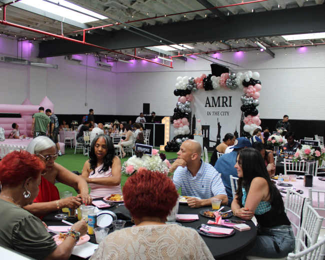 A group of people is dining at tables decorated with black and pink balloons in a spacious venue featuring a grassy area.