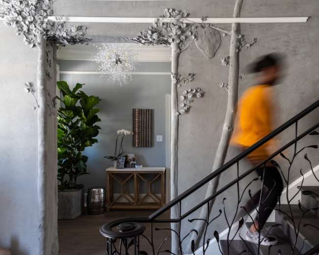 A person in a yellow jacket walks up a staircase in a modern entryway decorated with abstract tree-like wall sculptures and a large plant.