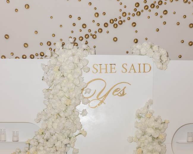 A table draped in a cream-colored cloth is set against a white wall adorned with artificial white flowers and the phrase "SHE SAID Yes" in gold lettering.