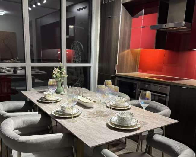 A modern dining area features a table set with plates and wine glasses, surrounded by upholstered chairs, against a backdrop of red and dark cabinetry.