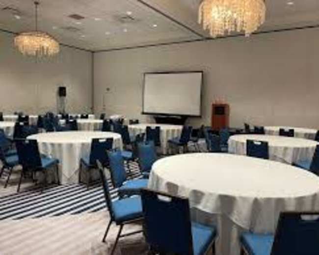 The image shows a conference room with several round tables covered in white tablecloths and blue chairs, along with a projector screen at the front.