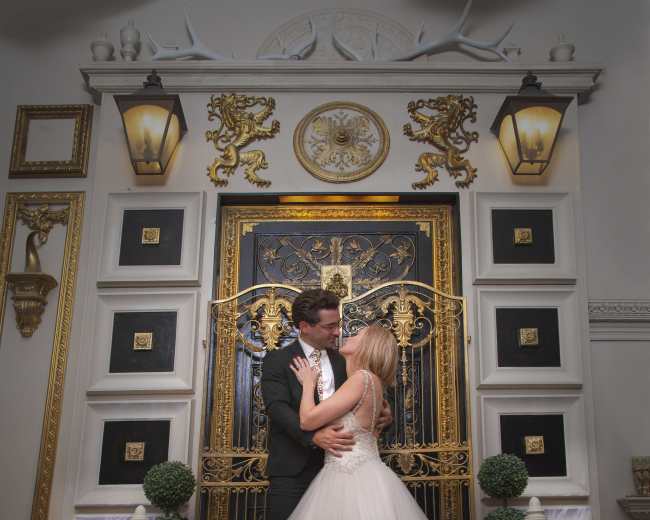 A couple embraces in front of an ornate gold and black decorative door within a lavishly styled interior.