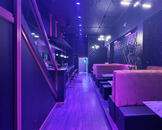 The image shows a modern interior of a dimly lit lounge with purple lighting, featuring plush seating and a bar area along one side.