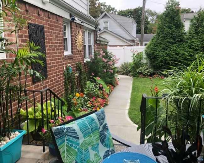A residential exterior with a pathway leading through a garden filled with colorful flowers, bordered by greenery and a brick wall.
