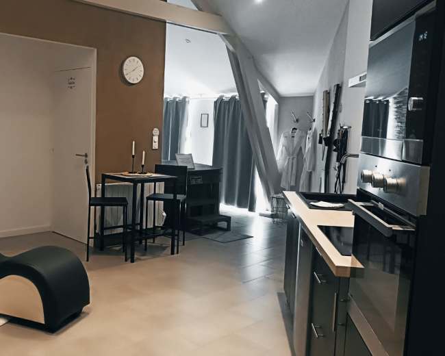 The image shows a modern interior space featuring a curved black lounge chair, a small dining table, and a kitchenette with built-in appliances.