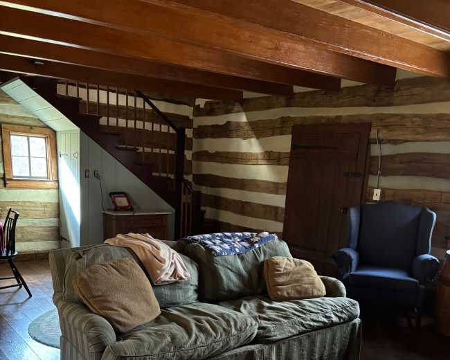 The image shows a cozy, rustic living room with a couch and chairs surrounded by wooden walls and a staircase in the background.