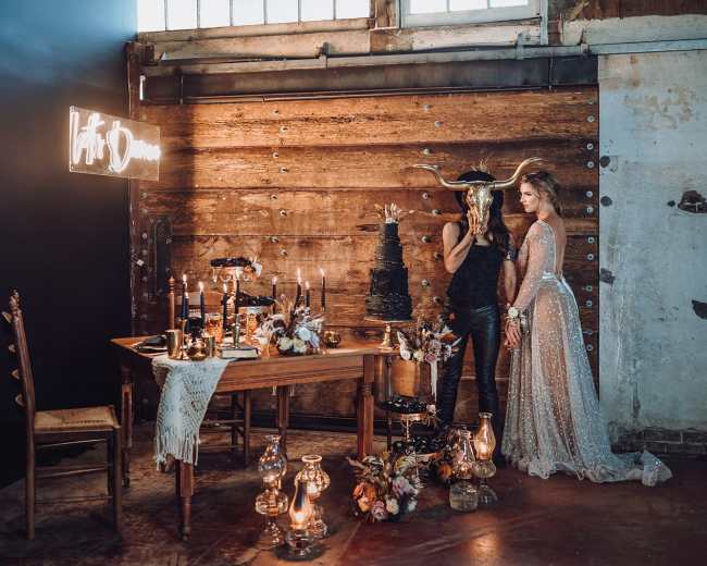 A decorated table with candles and floral arrangements is set against a rustic wooden backdrop, featuring a person holding a horned mask and another in a sparkly gown.