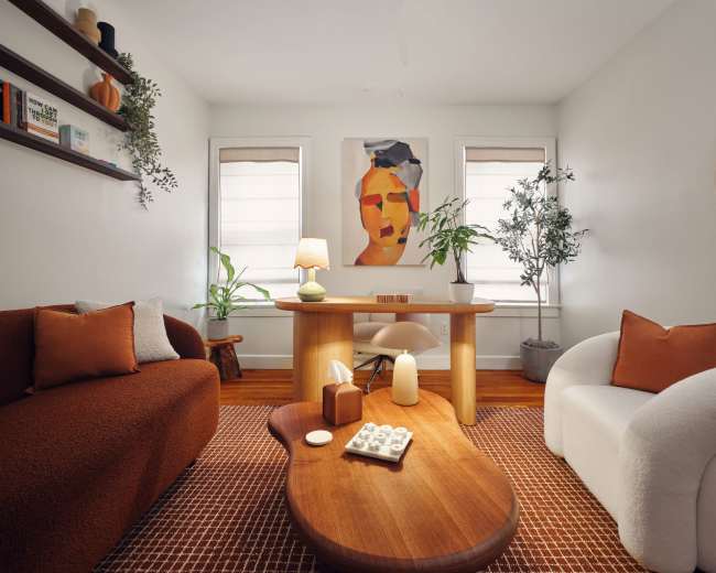 The image shows a cozy living room with two sofas, a wooden coffee table, a desk, and art on the walls, along with plants and decorative items.
